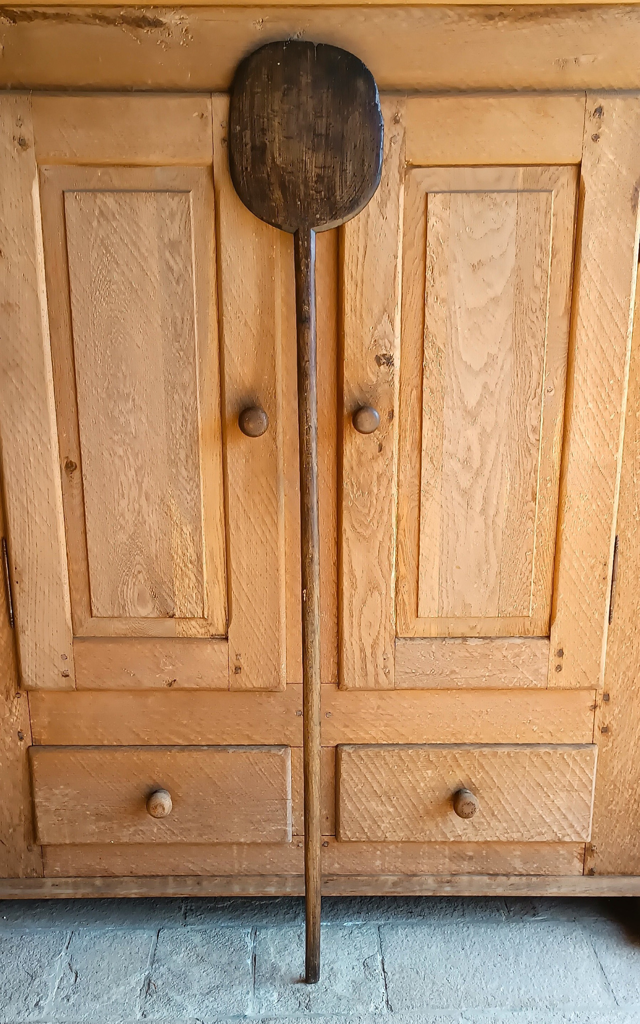 Antique Balkan wooden bread peel displayed vertically against a rustic wooden kitchen cabinet, showing its long handle and dark worn paddle
