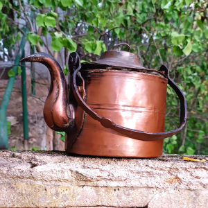 Antique copper kettle with lowered wrought iron bail handle, curved gooseneck spout, stepped lid and aged patina, photographed against green garden foliage.