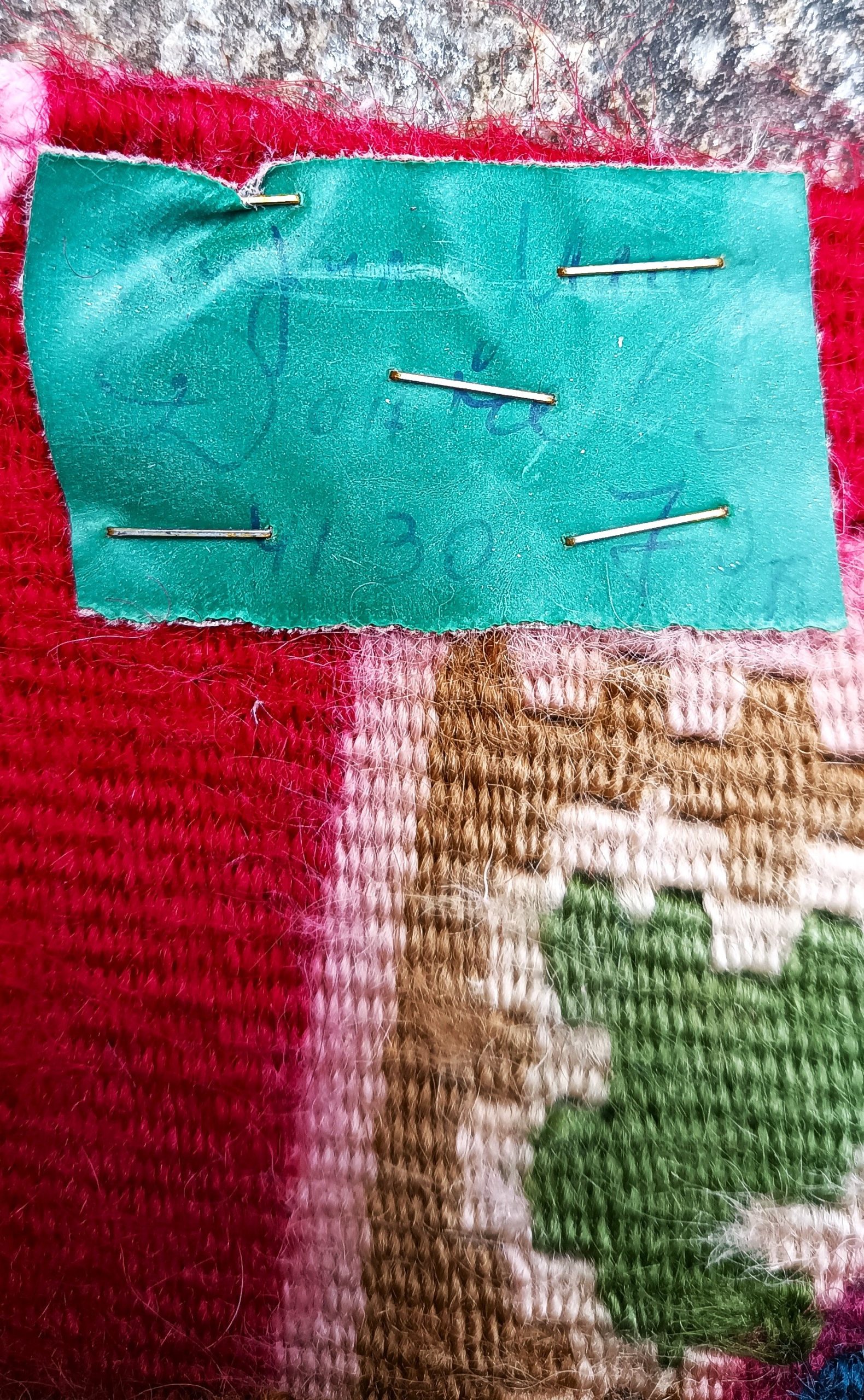 Faded green handwritten paper label attached with metal staples to the reverse of a traditional Kotel rug