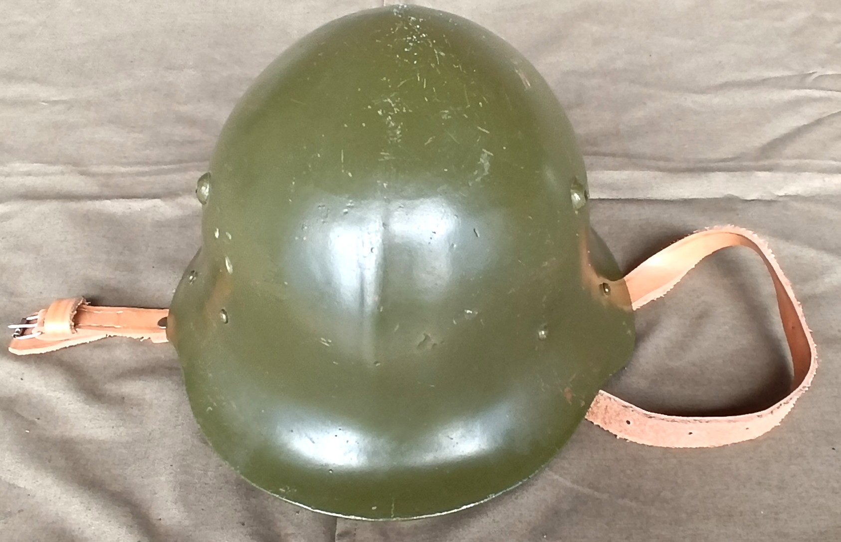 Top view of a Bulgarian M36 Type A steel helmet showing the central ridge, olive-green shell, retaining fasteners and leather chinstrap with buckle.