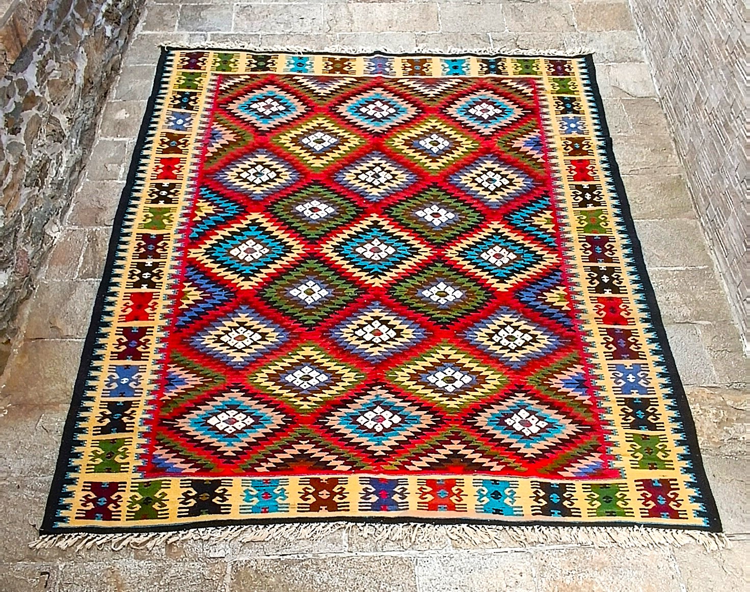 Full view of a Chiprovtsi Bakamski with Sofra rug with colourful stepped diamonds, white rosettes, cream geometric borders and fringes.
