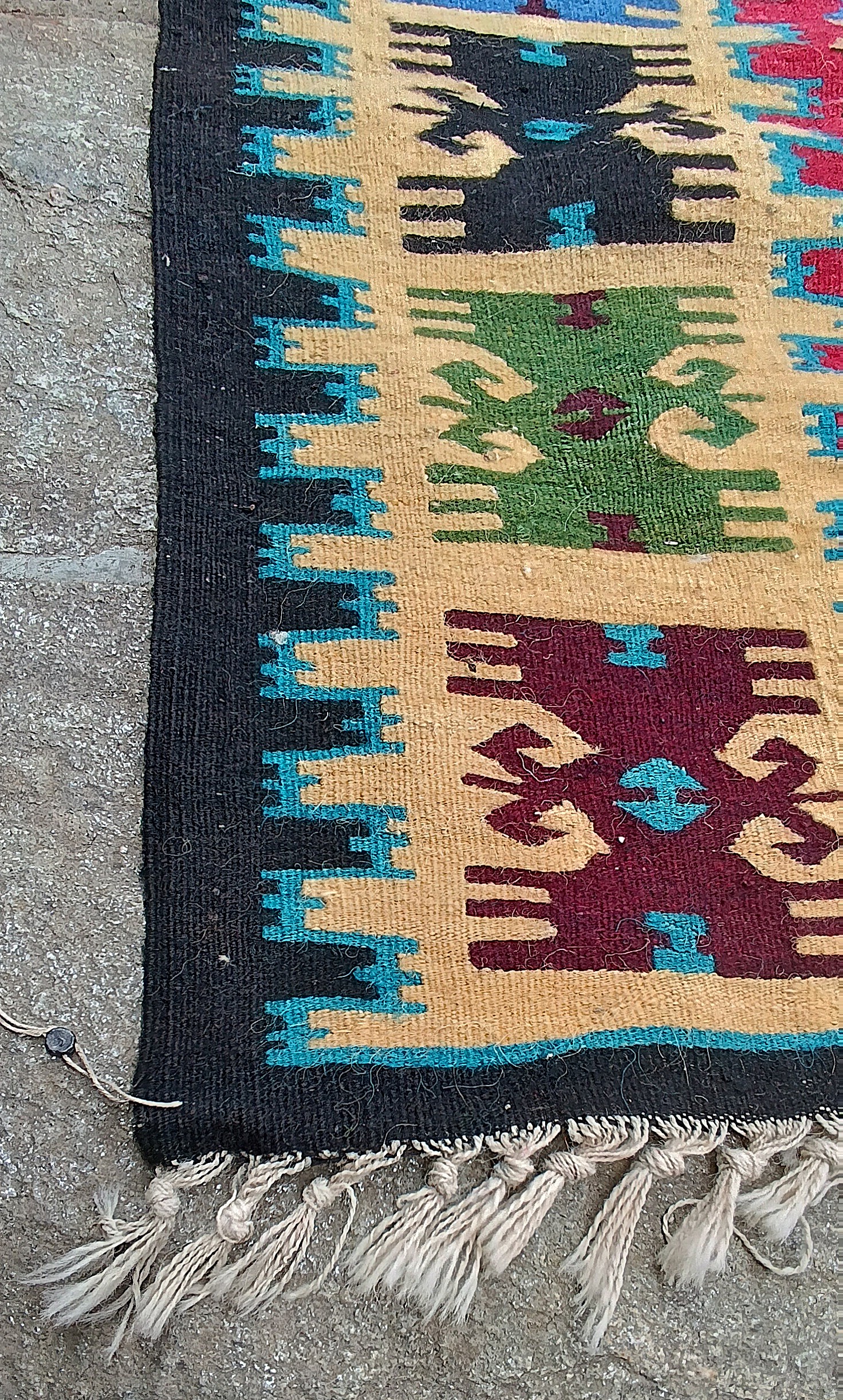Corner detail of a Chiprovtsi Bakamski rug with green and burgundy border ornaments, white fringes and a small attached seal.