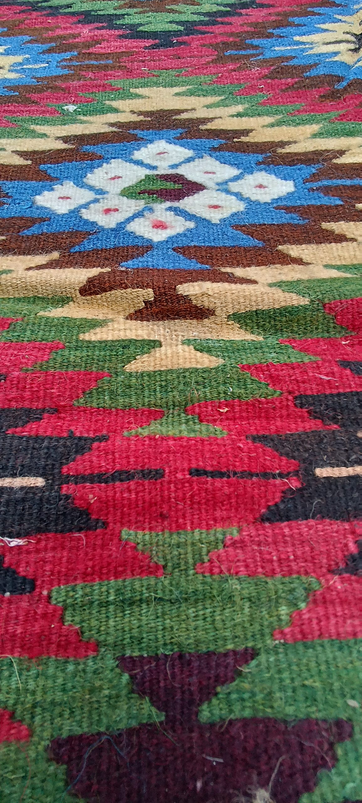 Low-angle wool texture close-up showing a white eight-part Sofra flower in blue with surrounding red, green and brown stepped shapes.