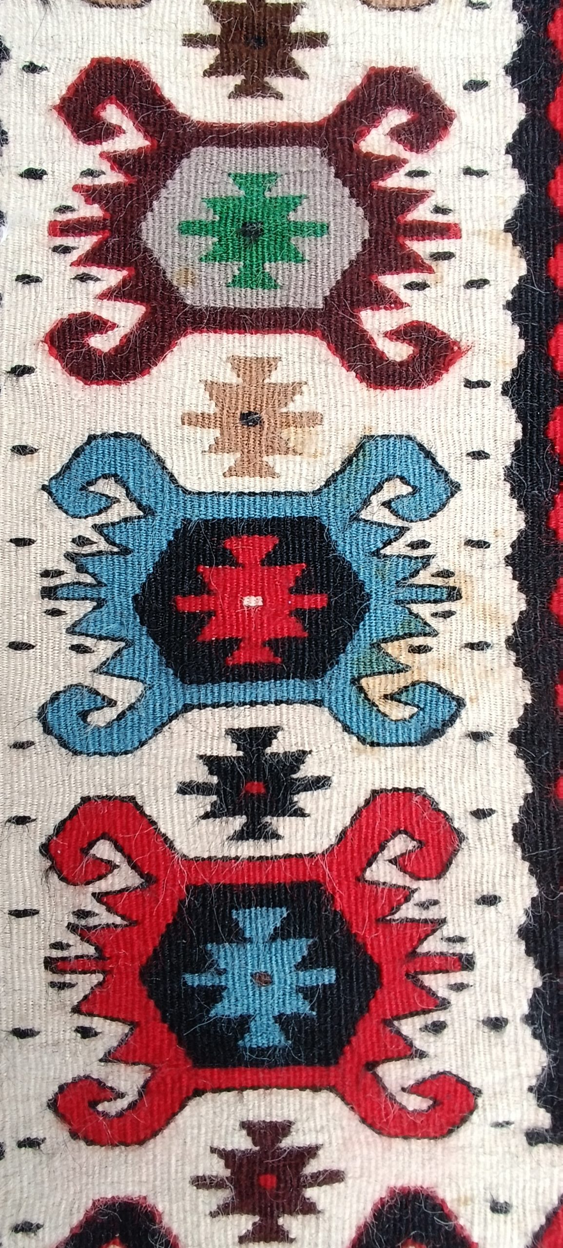 Macro photograph of the Chiprovtsi Bombichki rug border, showing blue, burgundy, green and brown figures separated by small cross-like forms.