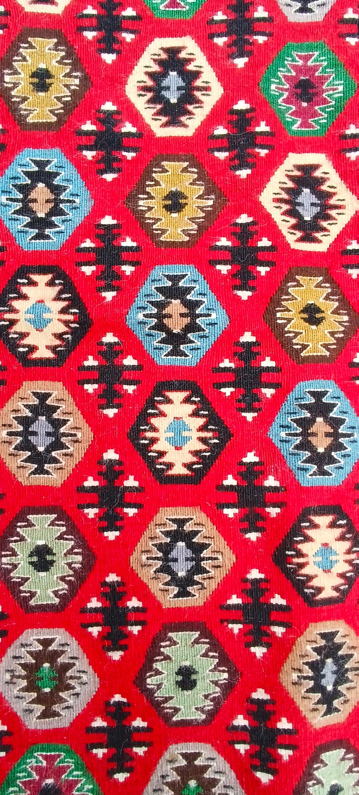 Detail of repeating stepped diamonds in blue, green, ochre, cream and black across the bright red field of a Bombichki rug.