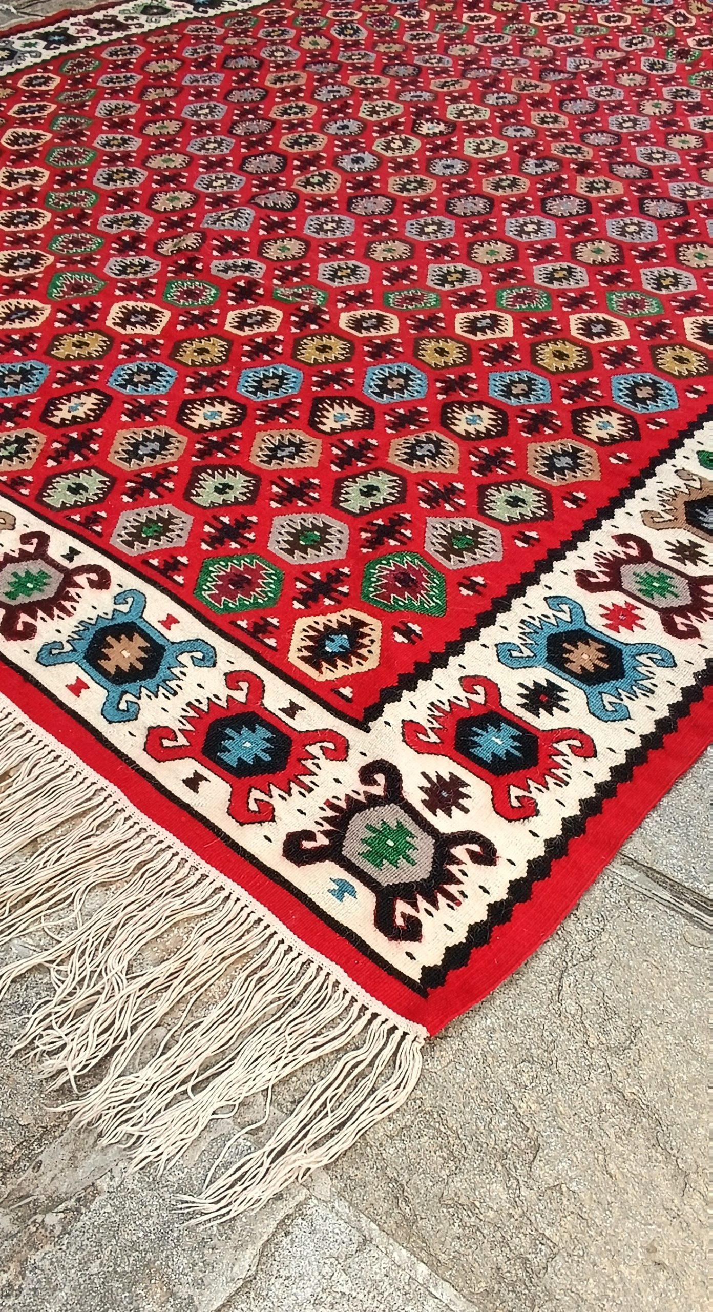 Corner of the Chiprovtsi Bombichki rug showing its red patterned field, cream geometric border and long white fringes.
