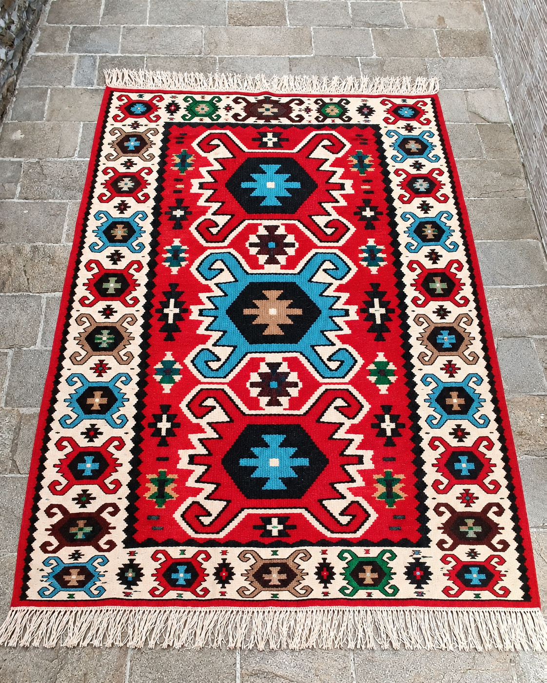 AI-generated artistic illustration of a red Bulgarian-style rug with three large Kanatitsa-inspired geometric motifs, turquoise and black accents, a cream border and white fringes.