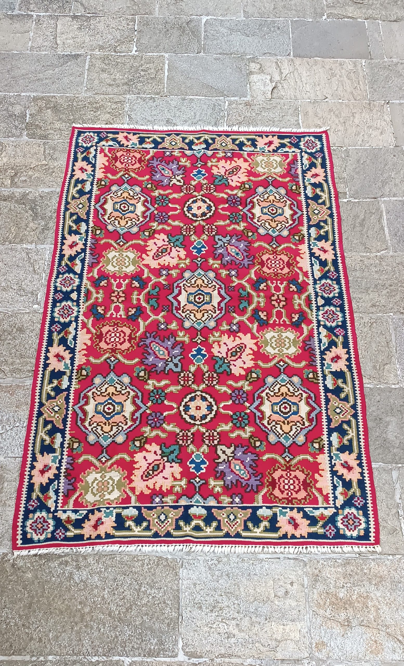 Traditional red rug with colourful floral rosettes, symmetrical ornamental medallions, a dark blue floral border and short light-coloured fringes.