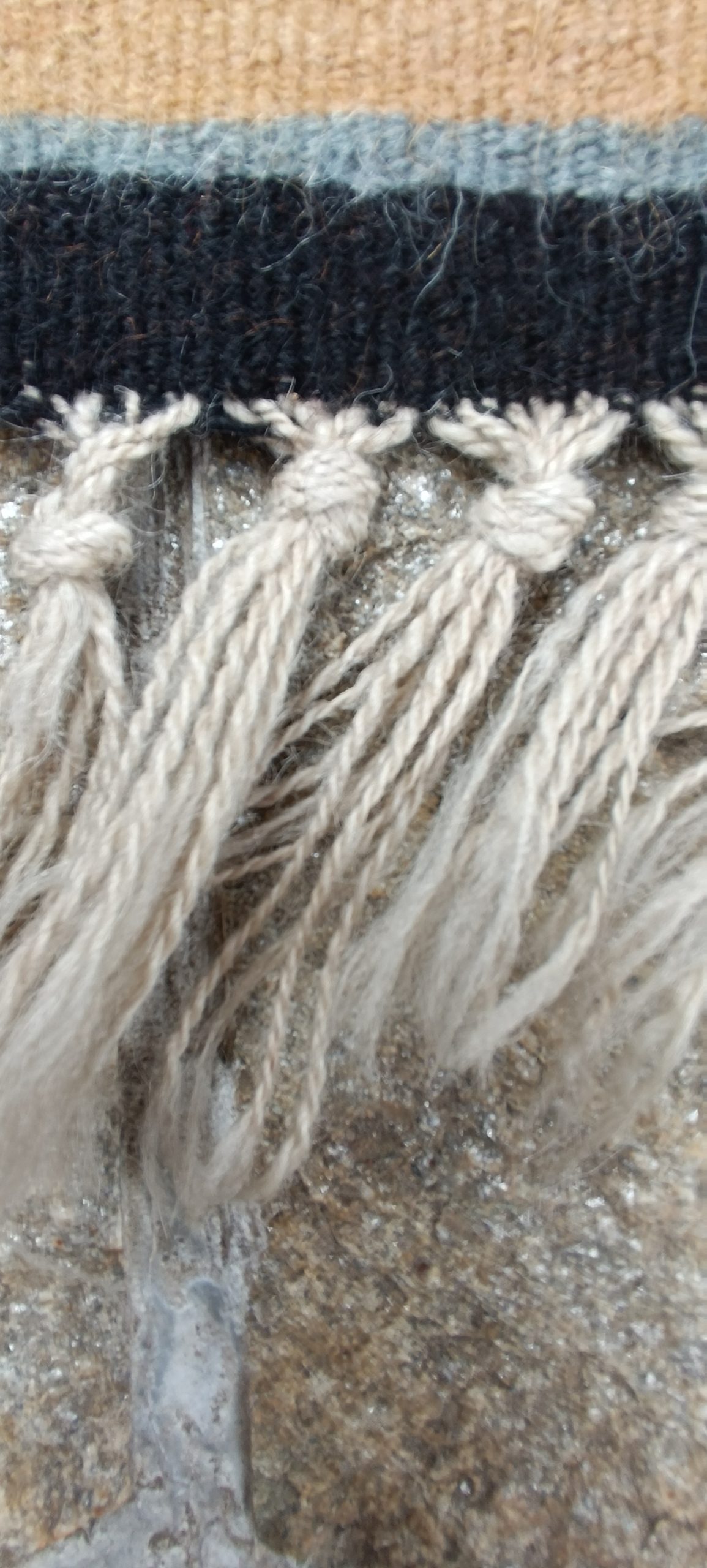 Close-up of twisted and knotted white wool warp fringes along the dark woven edge of an old Bulgarian Chiprovtsi rug.
