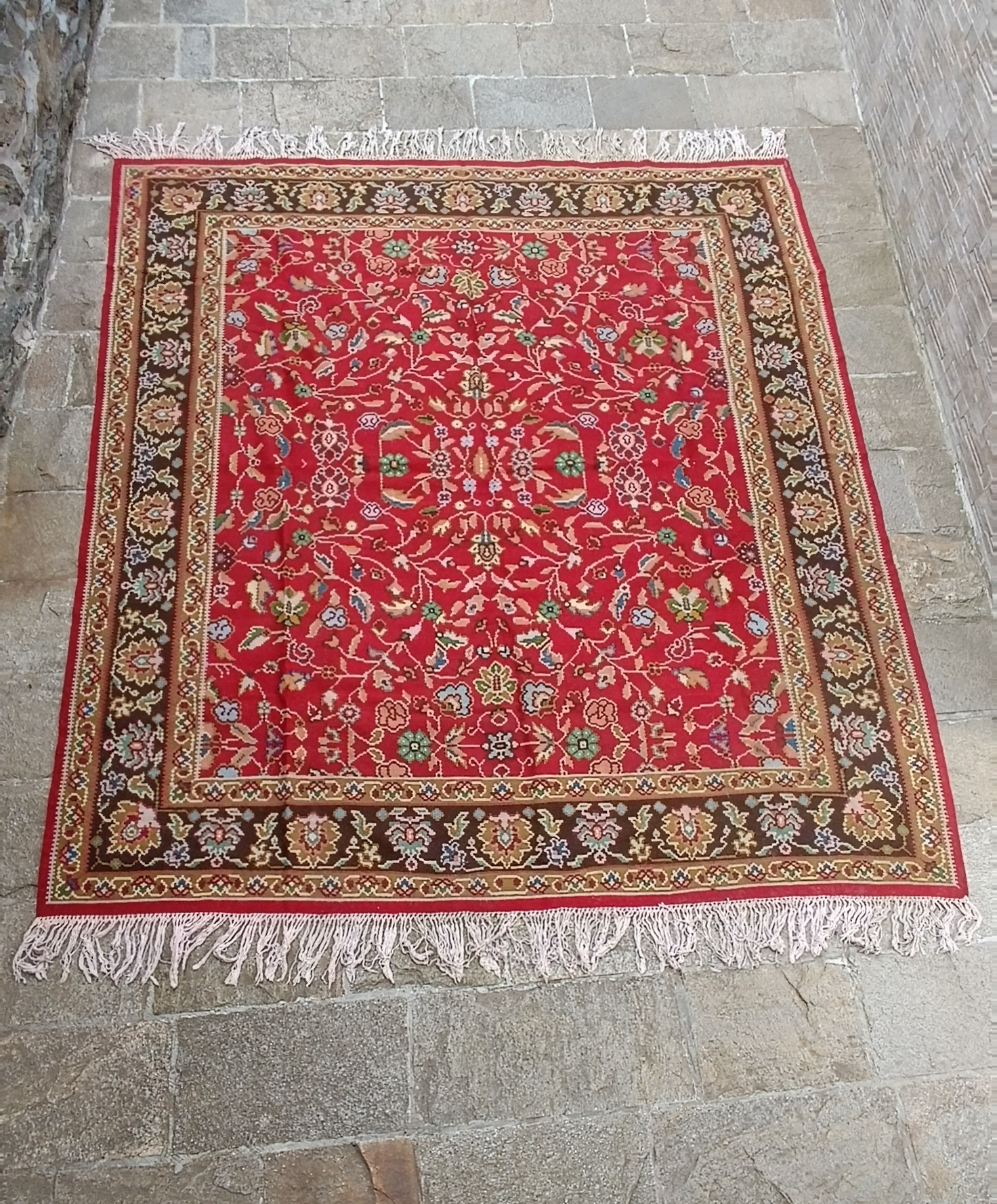 Traditional red rug with intricate symmetrical floral vines, a broad dark brown flower-patterned border and long cream-coloured fringes.