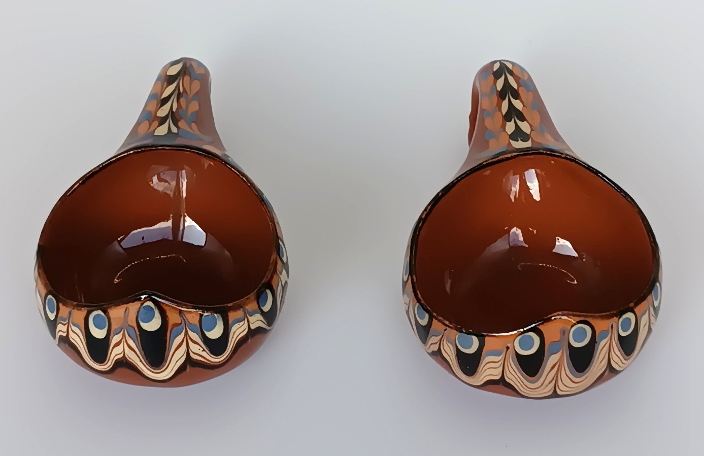 Top view of two vintage Bulgarian Troyan pottery miniature cups with glossy brown interiors, curved handles and traditional Peacock Eye decoration in blue, cream and black.
