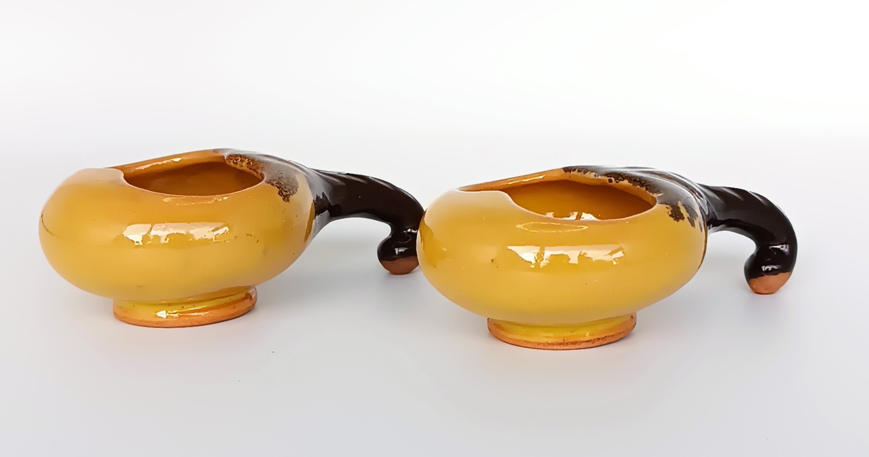 Side view of a pair of vintage ceramic Schnapspfeifle pipe-shaped liqueur cups with ochre yellow bodies and dark brown curved handles