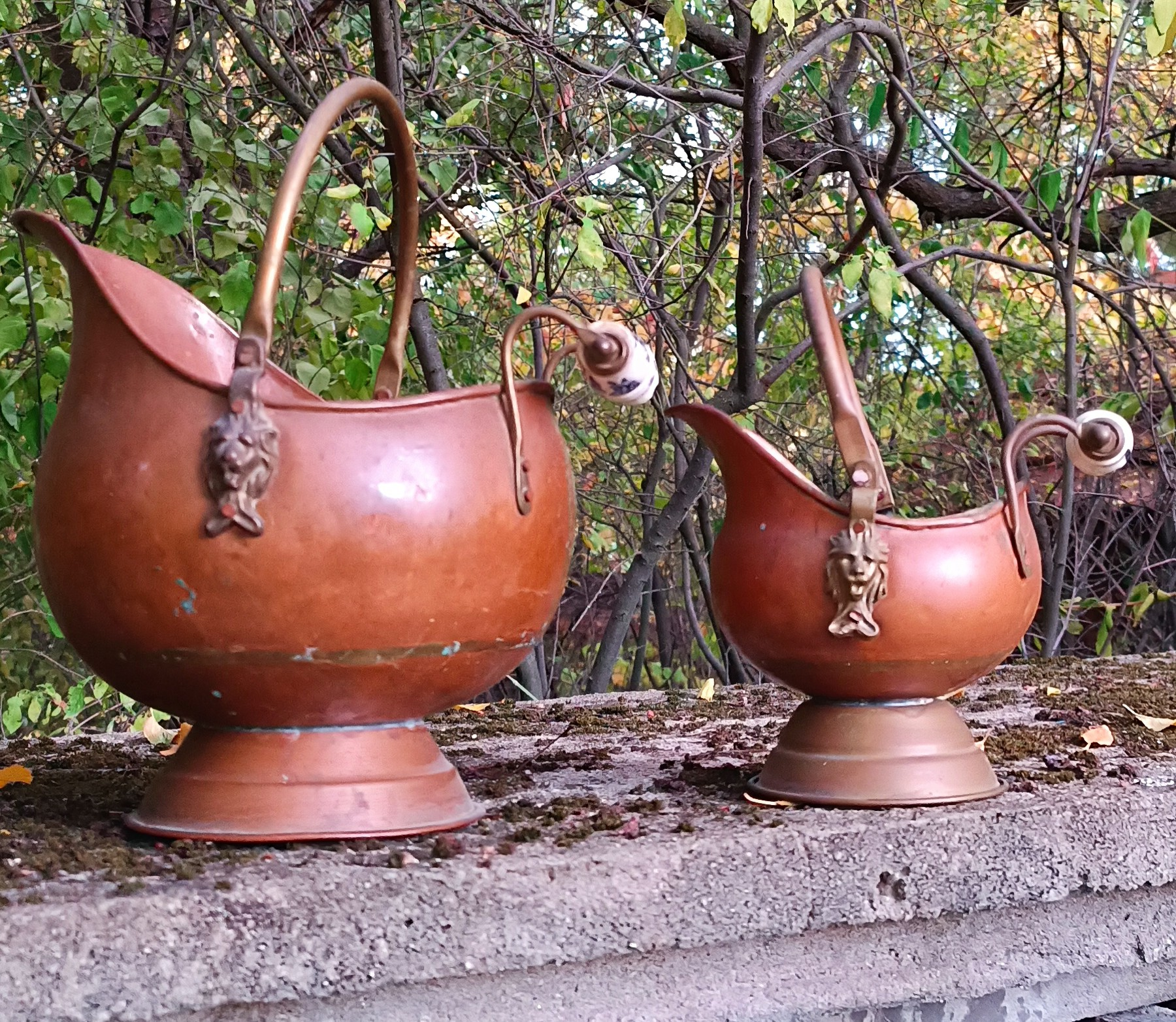 Front view of two vintage Dutch copper coal scuttles in different sizes, with decorative lion-head mounts, arched handles, Delft-style ceramic grips and pedestal bases.