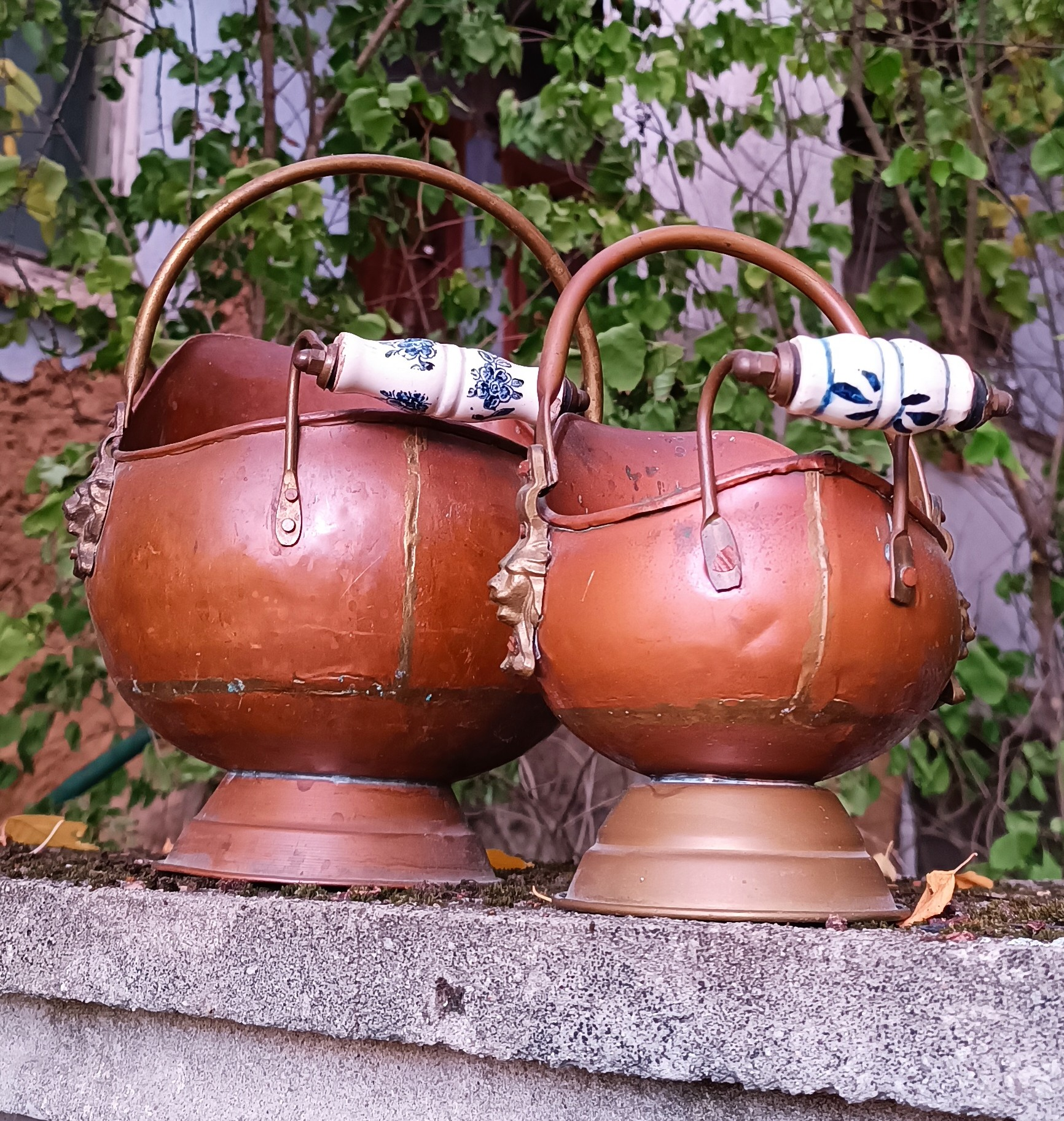 Rear view of two vintage Dutch copper coal scuttles with blue-and-white Delft-style ceramic handles, arched metal grips, riveted fittings, visible construction seams and pedestal bases.