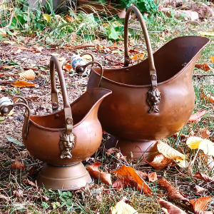Set of two vintage Dutch copper coal scuttles in different sizes, featuring lion-head mounts, curved metal handles, Delft-style blue-and-white ceramic grips and pedestal bases.