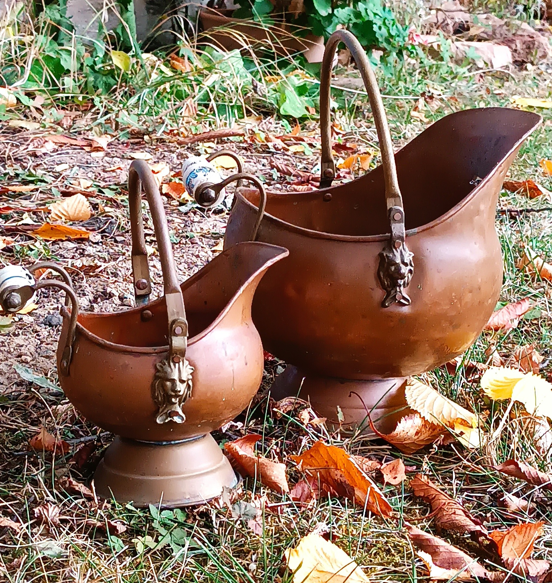 Set of two vintage Dutch copper coal scuttles in different sizes, featuring lion-head mounts, curved metal handles, Delft-style blue-and-white ceramic grips and pedestal bases.