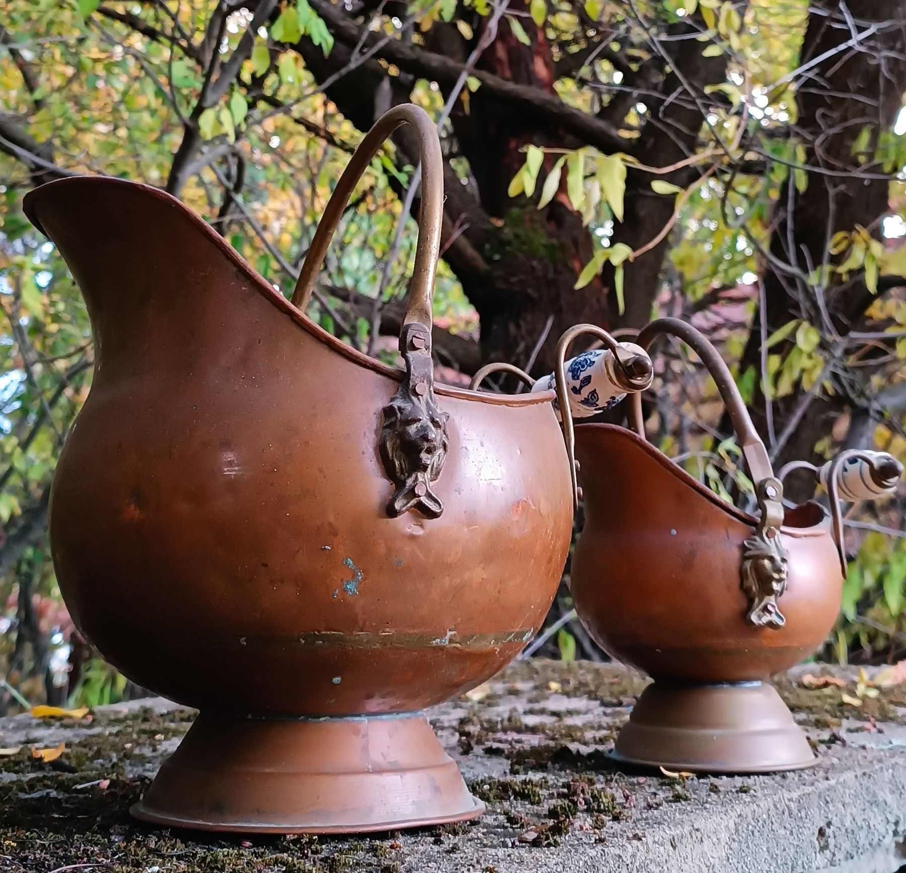Side view of two vintage Dutch copper coal scuttles in different sizes with lion masks, metal handles and Delft-style ceramic grips