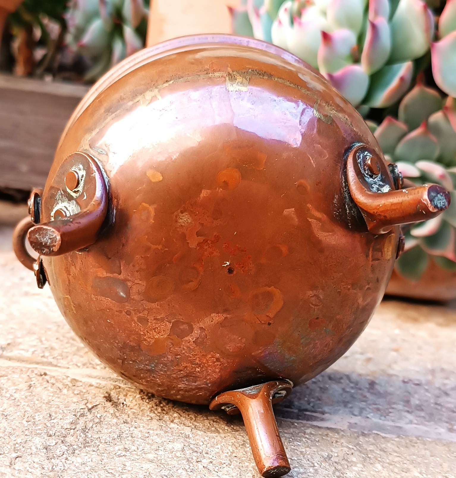 Underside of vintage three-legged copper cauldron showing rounded base, riveted foot attachments and uneven oxidation patina
