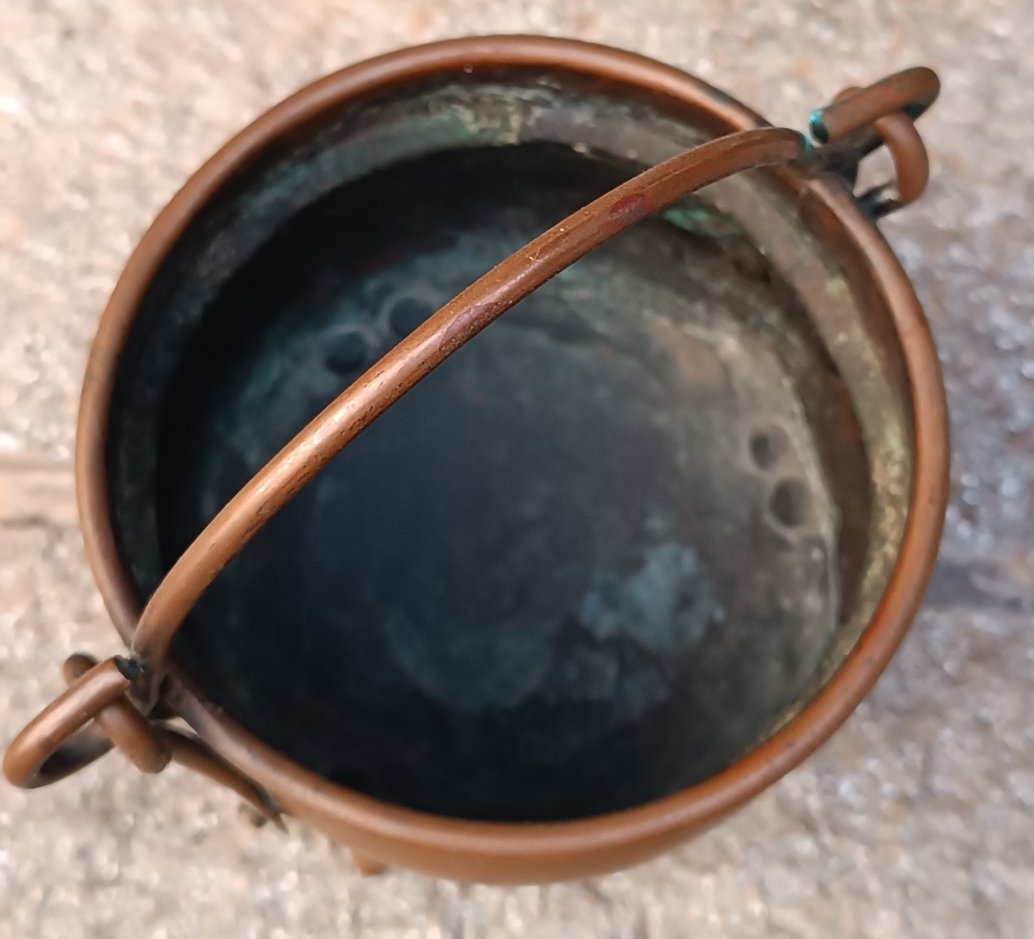 Top view inside a vintage three-legged copper cauldron, showing its dark oxidised interior, greenish patina, rolled copper rim and movable arched carrying handle.