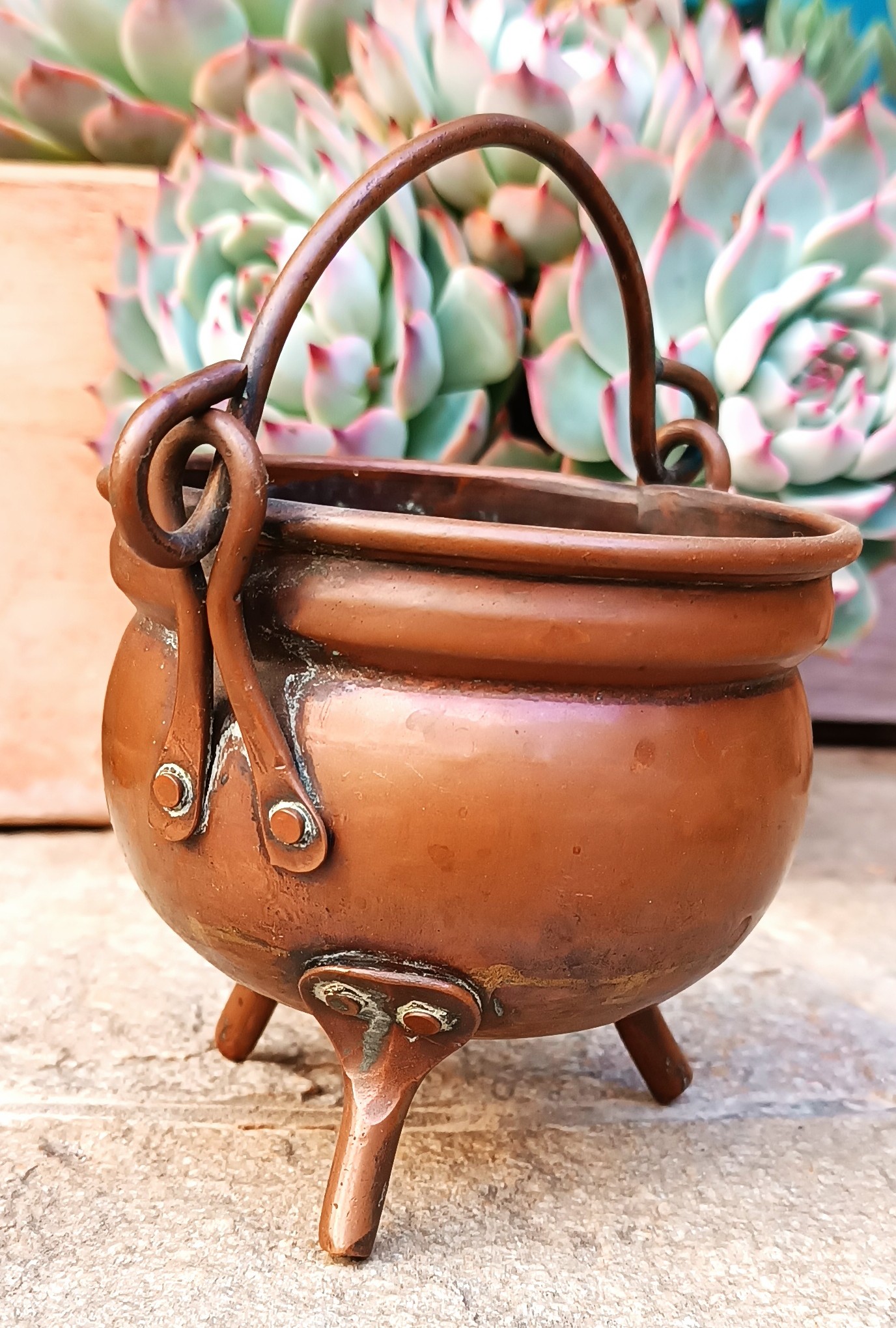 Side view of vintage copper cauldron showing three riveted legs, rounded metal body, side fittings and swing handle