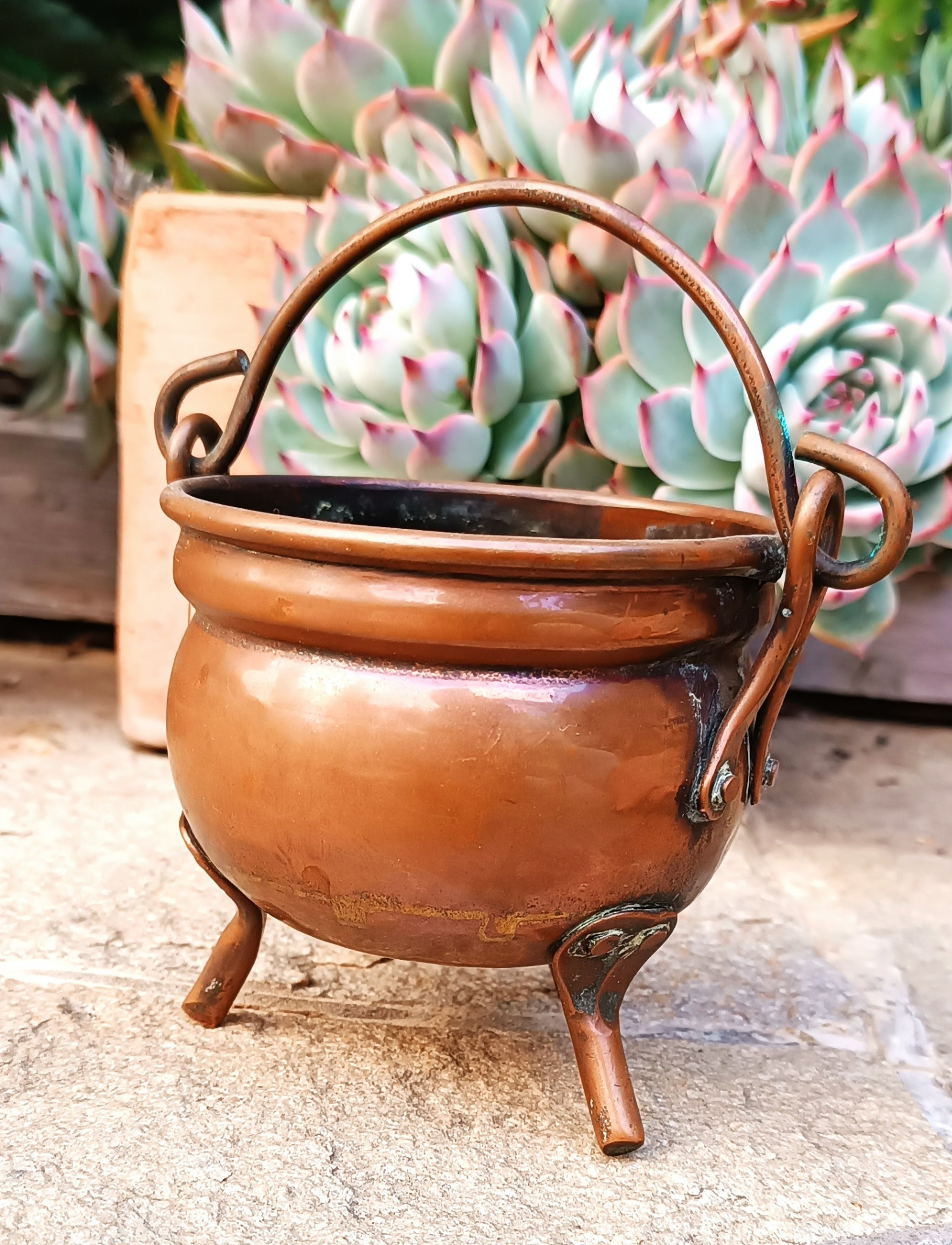 Side view of a vintage three-legged copper cauldron with a rounded body, raised rolled rim, movable arched handle, riveted metal fittings and natural aged patina.