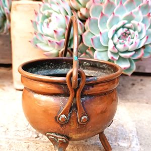 Vintage three-legged copper cauldron with rounded body, riveted metal legs, arched swing handle and aged patina, photographed against decorative succulent plants.