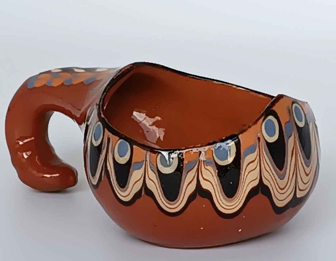 Close-up of a vintage Bulgarian Troyan pottery miniature cup with a curved handle, terracotta-brown glaze and traditional Peacock Eye decoration in cream, black and blue.