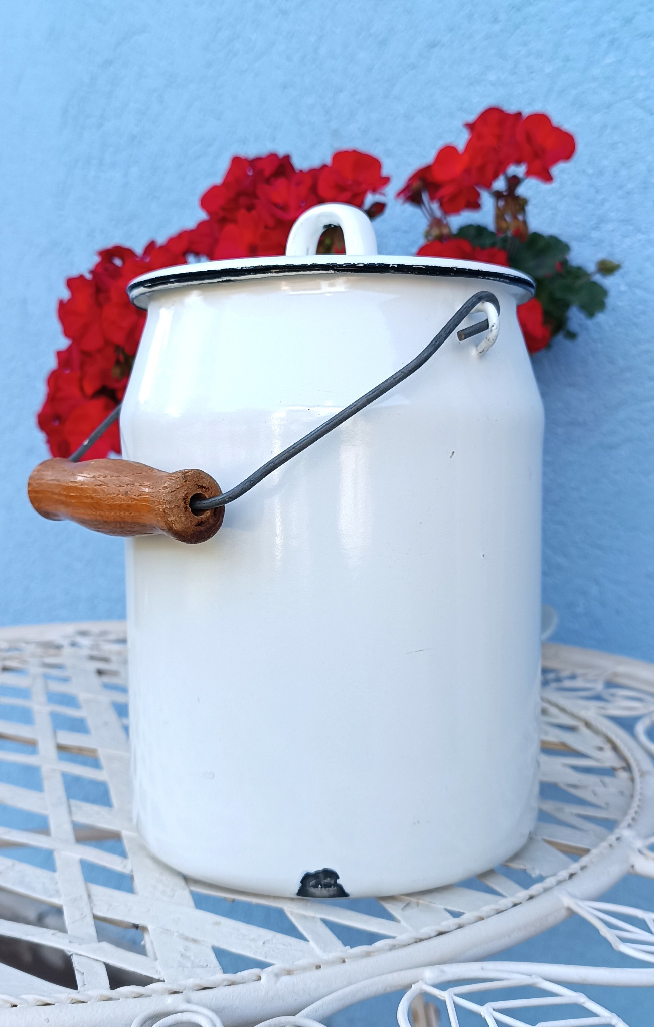 Front view of a vintage white enamel milk pail with lid, black rim and wooden carrying handle, showing a chipped area near the base.