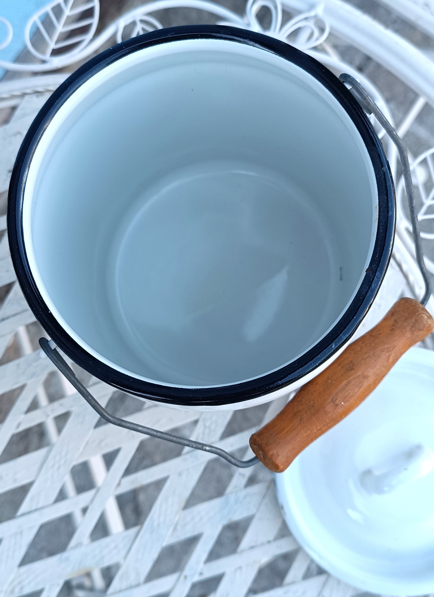 Top view inside a vintage white enamel milk pail showing its clean-looking interior, black rim, metal carrying handle, wooden grip and removable lid.