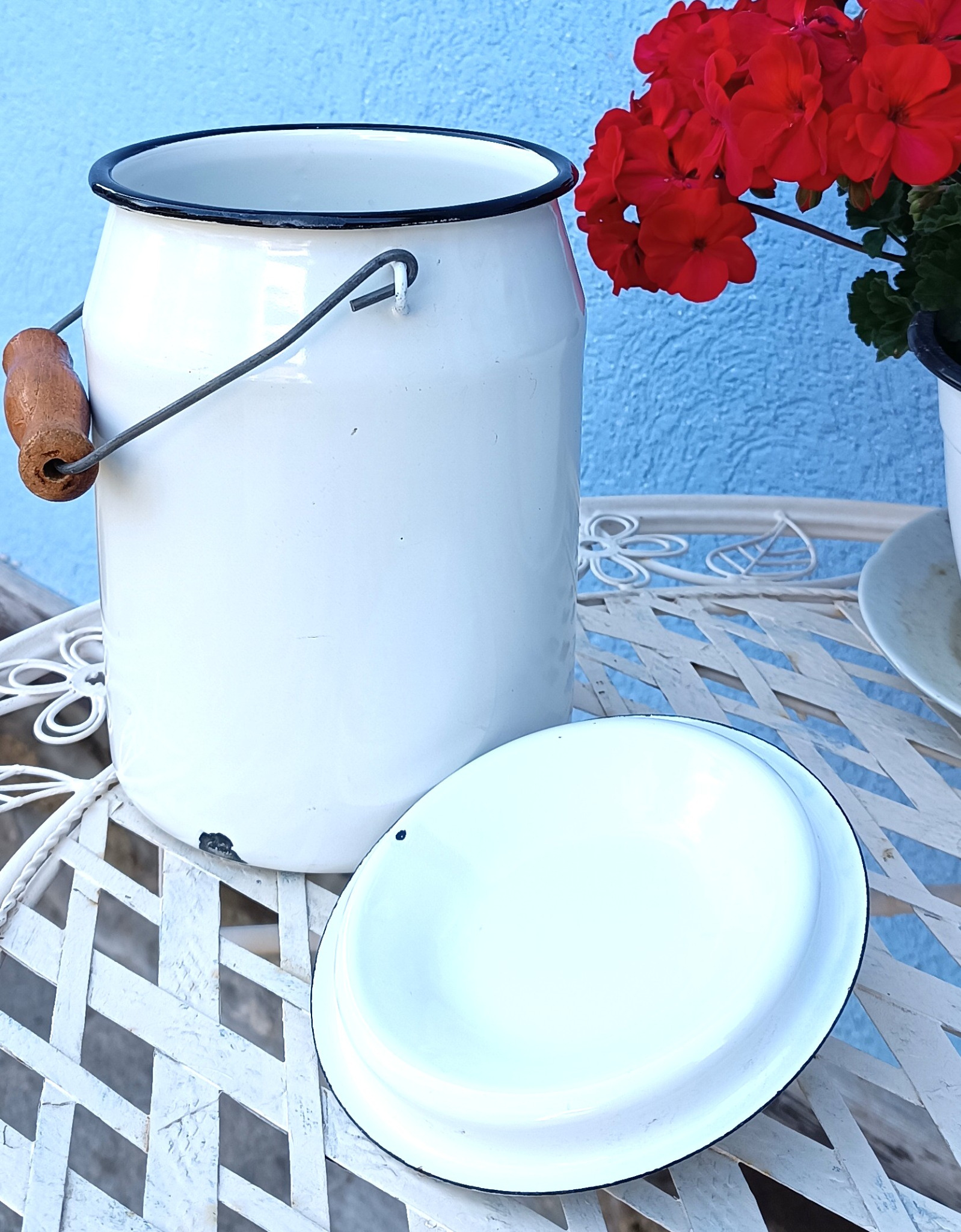 Vintage white enamel milk pail with black rim and wooden carrying handle, displayed open beside its removable lid on a garden table with red geraniums.